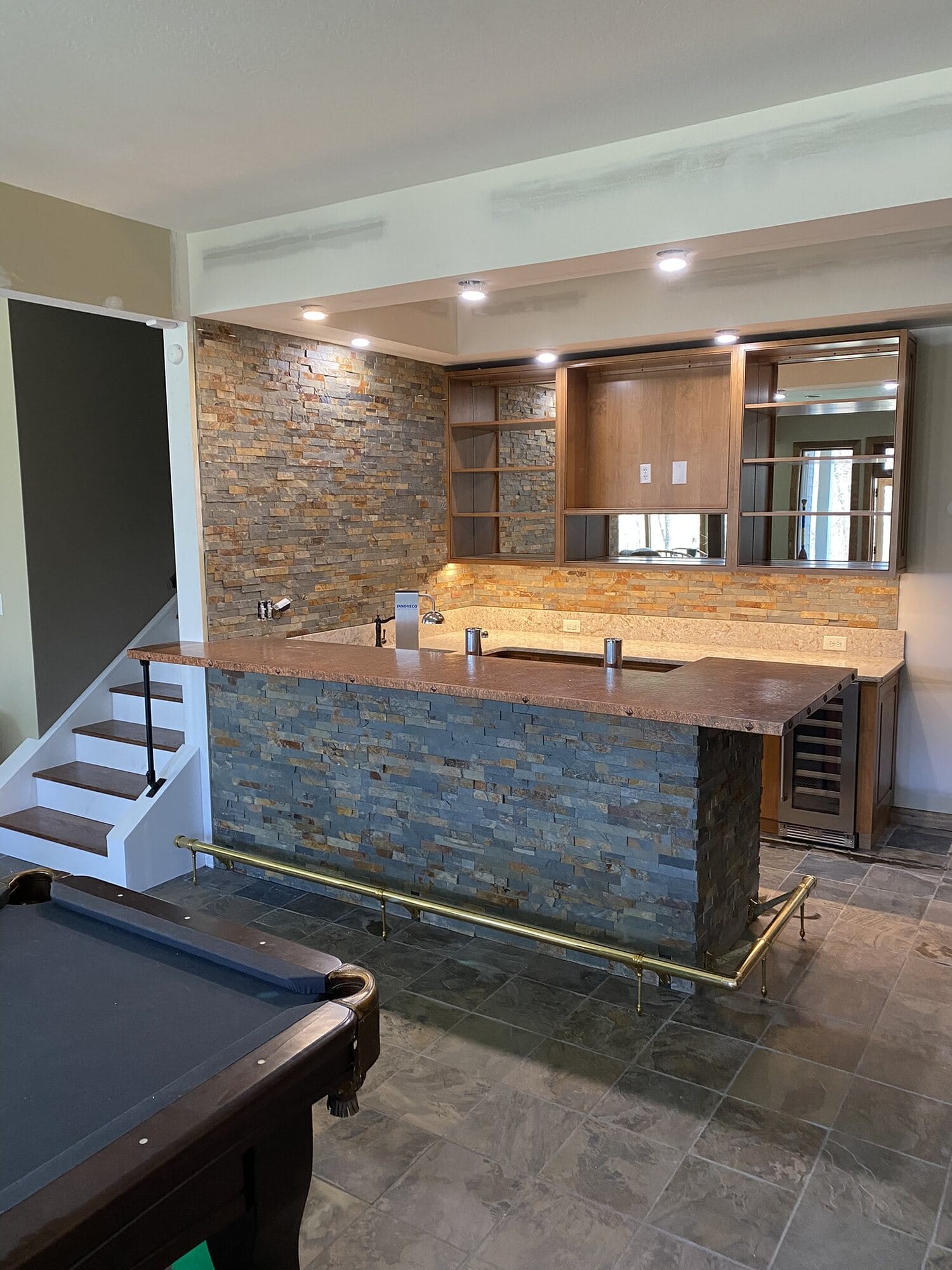 Finished basement bar with a stacked stone bar front, wood countertop, foot rail, wine storage, and adjacent pool table.