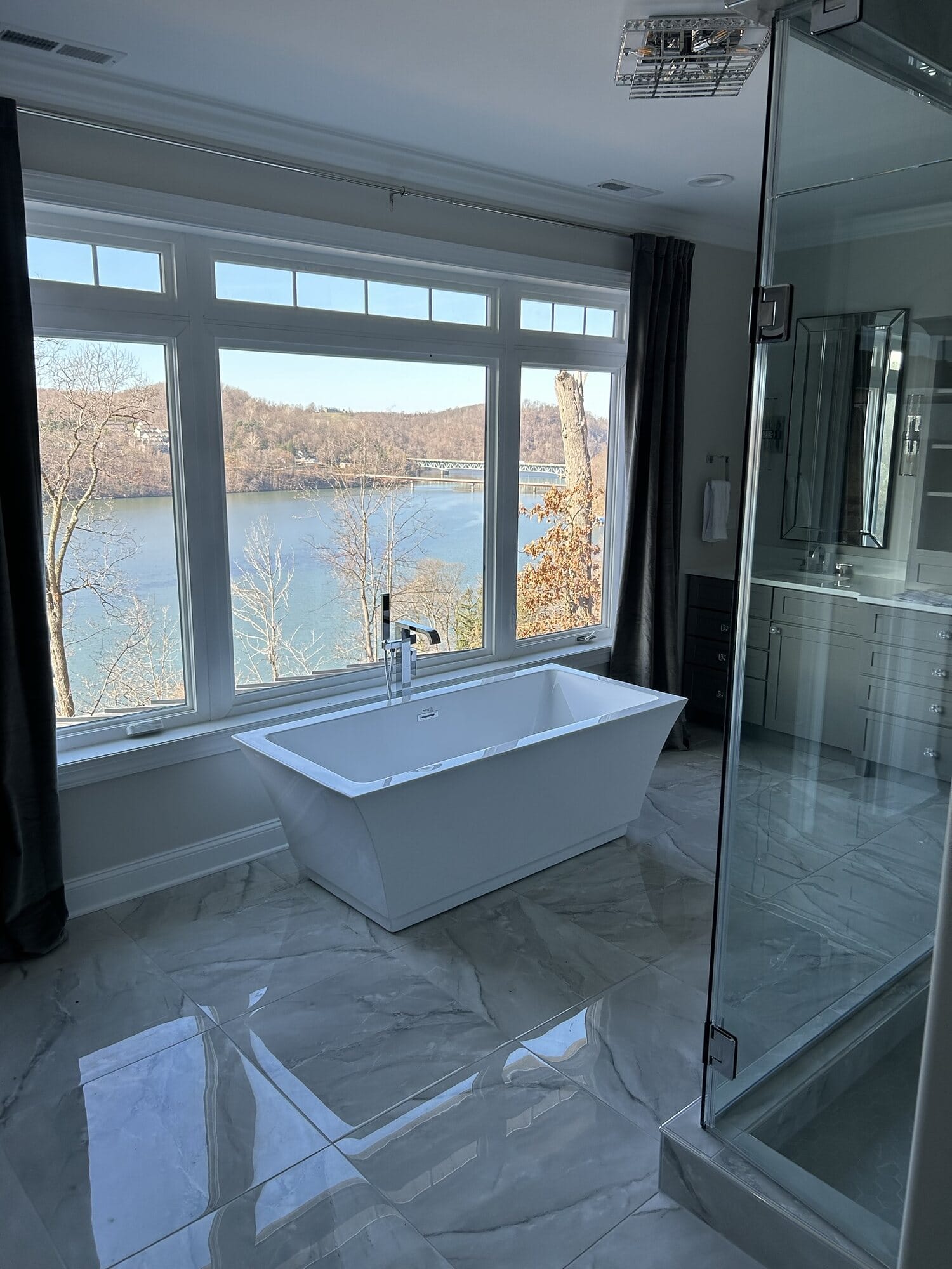 Spa-style bathroom with a freestanding soaking tub set before a picture window, frameless glass shower, and large-format gray tile.