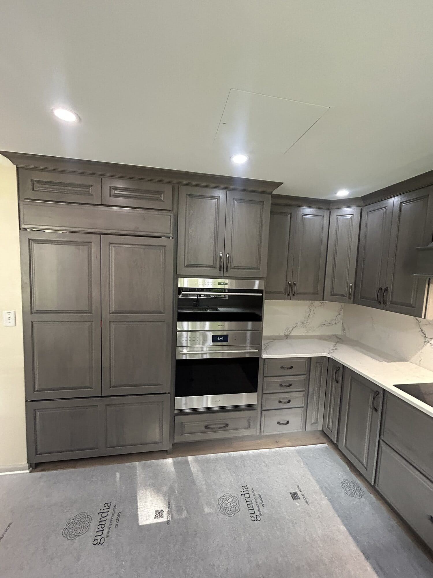 Gray stained kitchen corner with tall pantry cabinetry, stacked wall ovens, and marble-look quartz countertops.