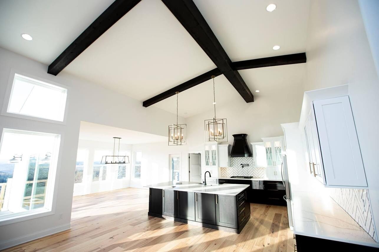 Open great room kitchen with a vaulted ceiling, exposed black beams, white cabinetry, dark island, and glass pendant lighting.