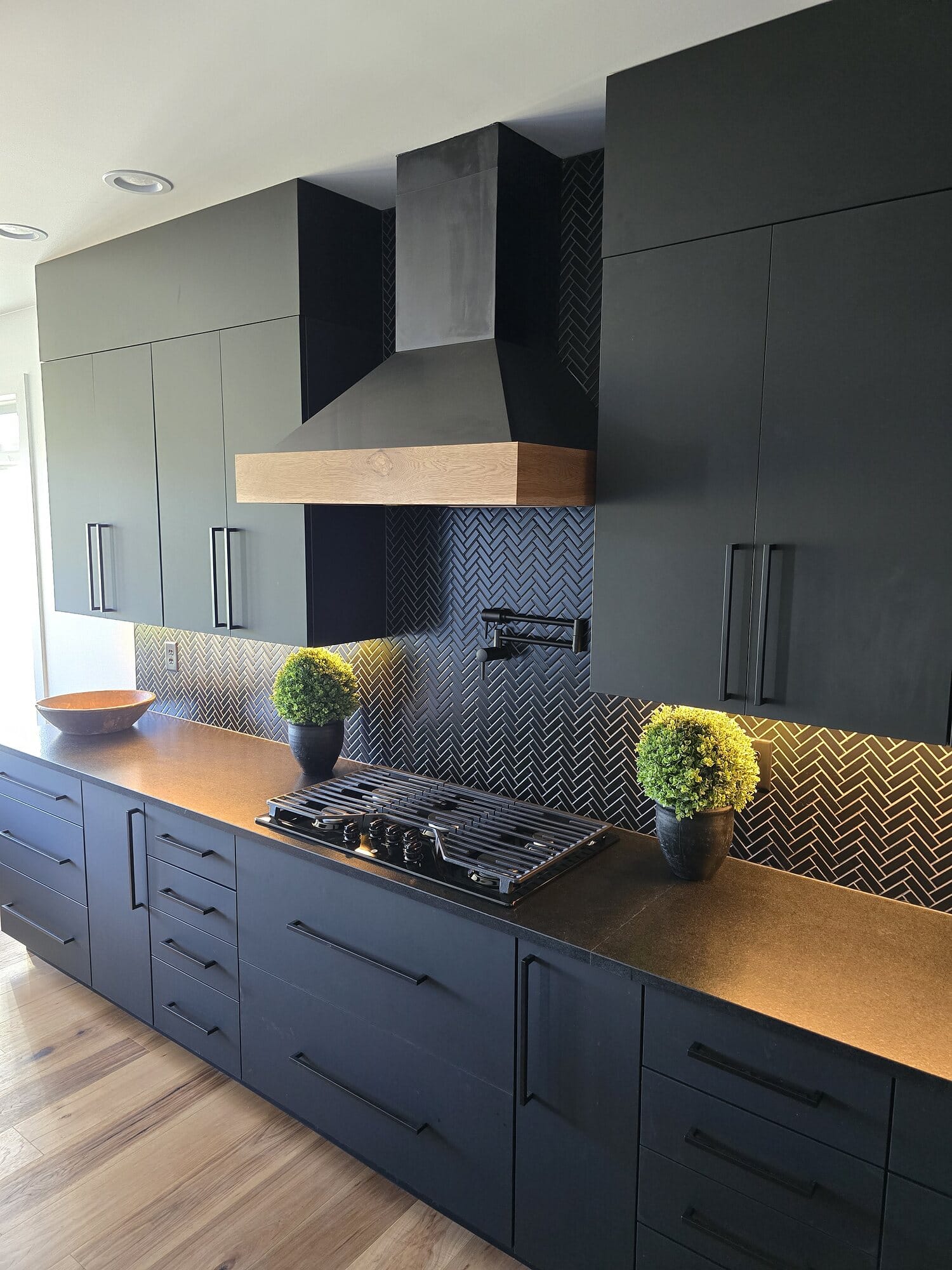 Modern kitchen remodel with floor-to-ceiling black cabinetry, a white oak hood mantel, gas cooktop, and a dark herringbone tile backsplash.