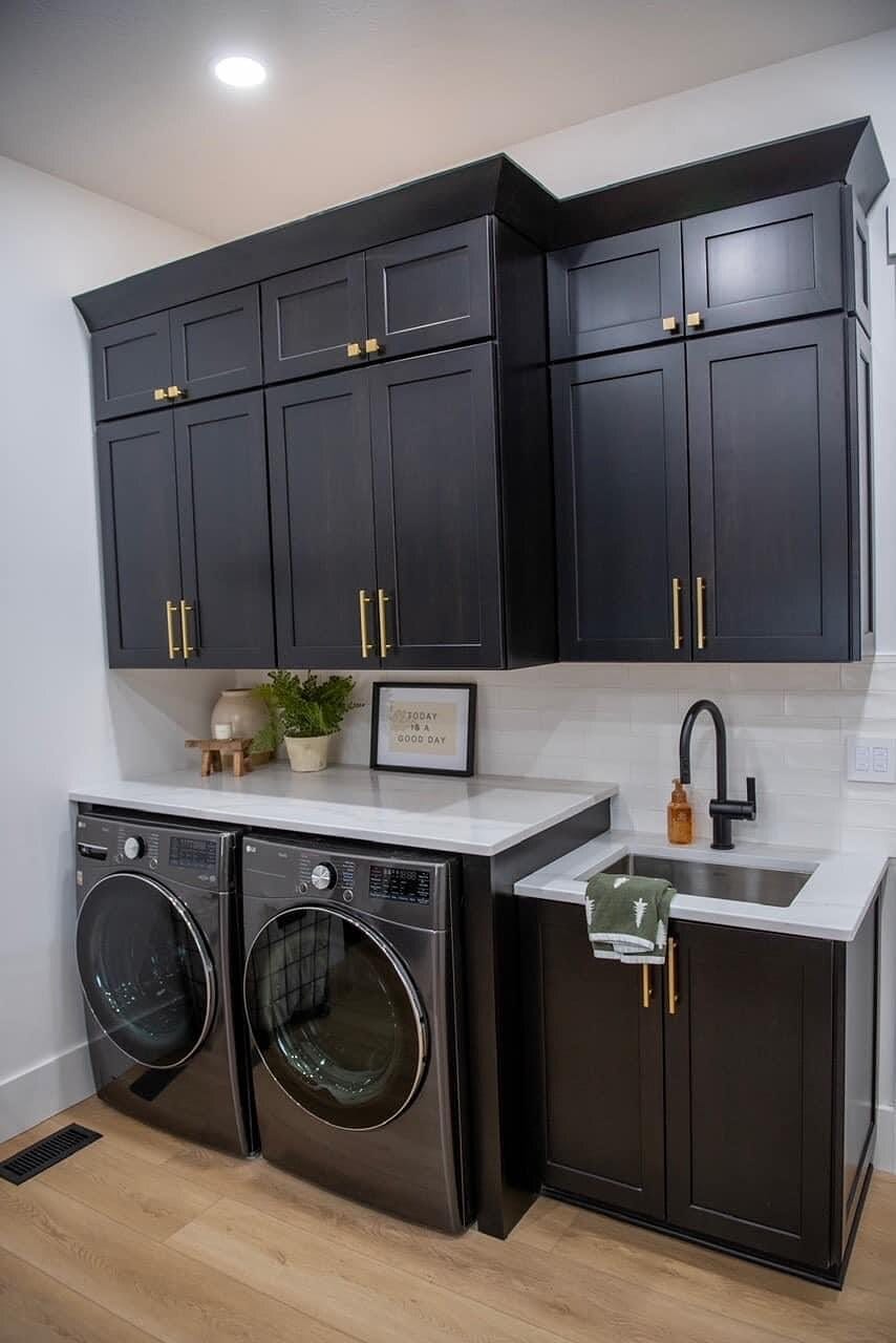 Laundry room with navy cabinetry, brushed gold hardware, quartz folding counter, undermount sink, and front-load appliances.
