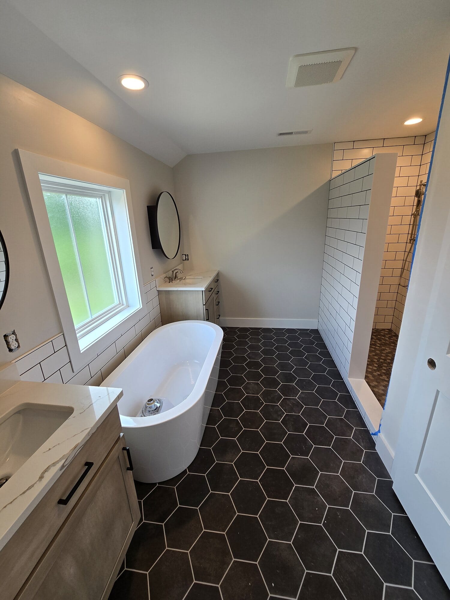 Master bathroom remodel with a freestanding soaking tub, black hexagon floor tile, white subway tile walls, round mirror, and a floating wood vanity.