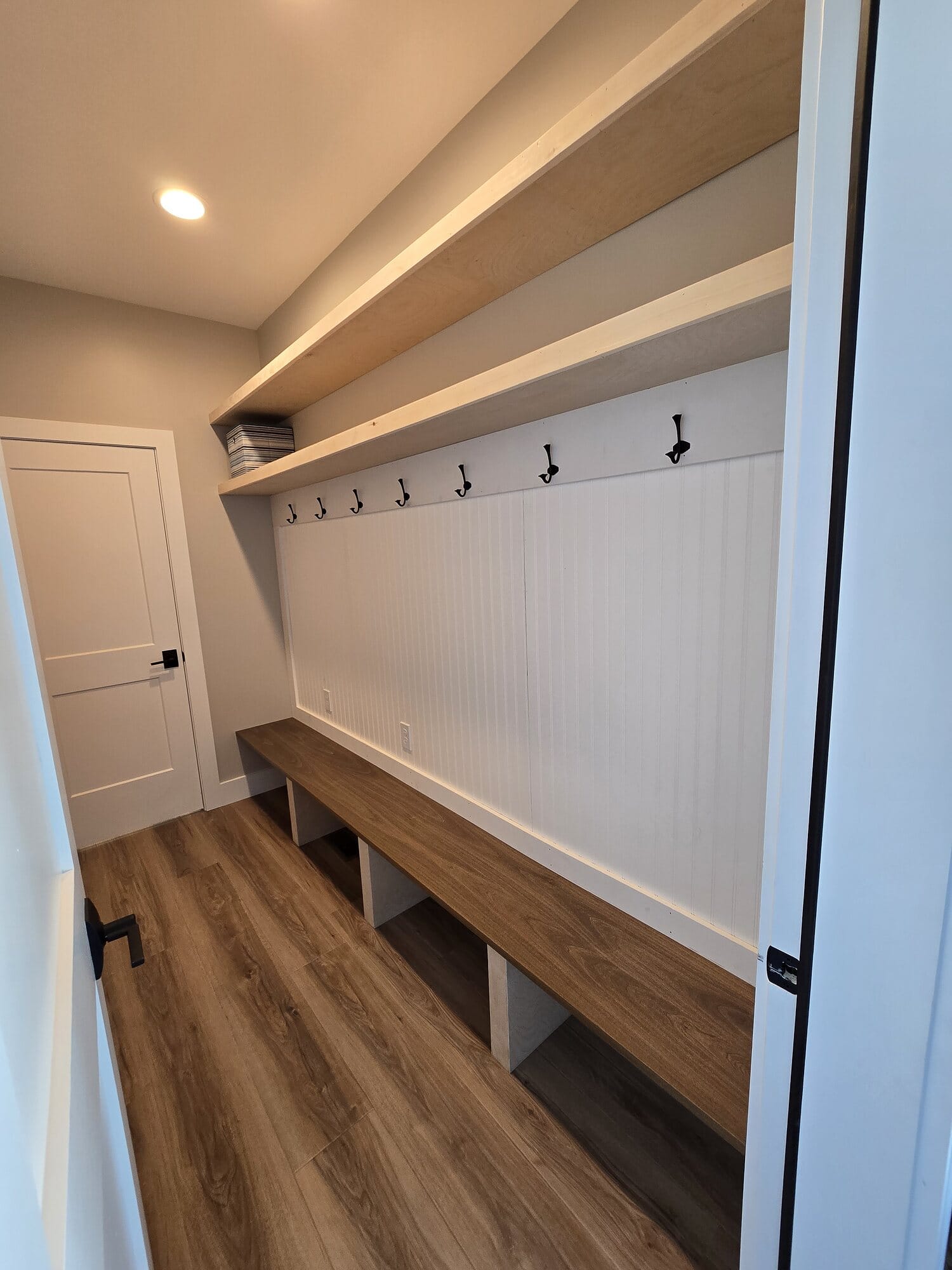 Built-in mudroom bench with open cubby storage, upper shelf, coat hooks, and white beadboard wainscot over wide-plank wood flooring.