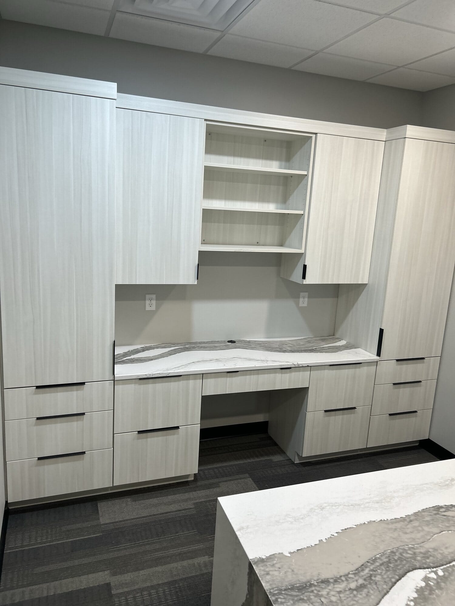 Custom office built-in with light woodgrain slab cabinetry, open shelving, integrated desk, and a veined quartz countertop.