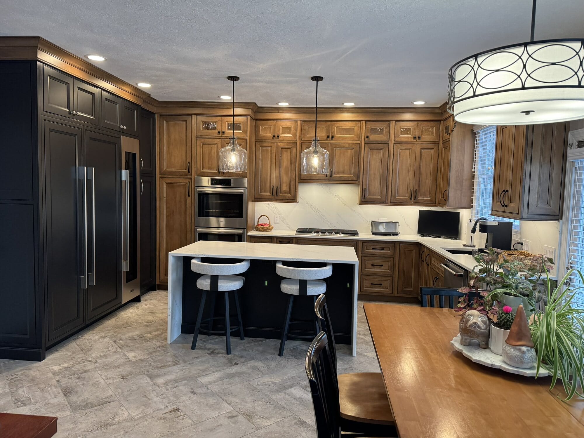 Wide view of a two-tone kitchen remodel with stained wood cabinetry, black tall cabinets, island seating, and a built-in desk area.