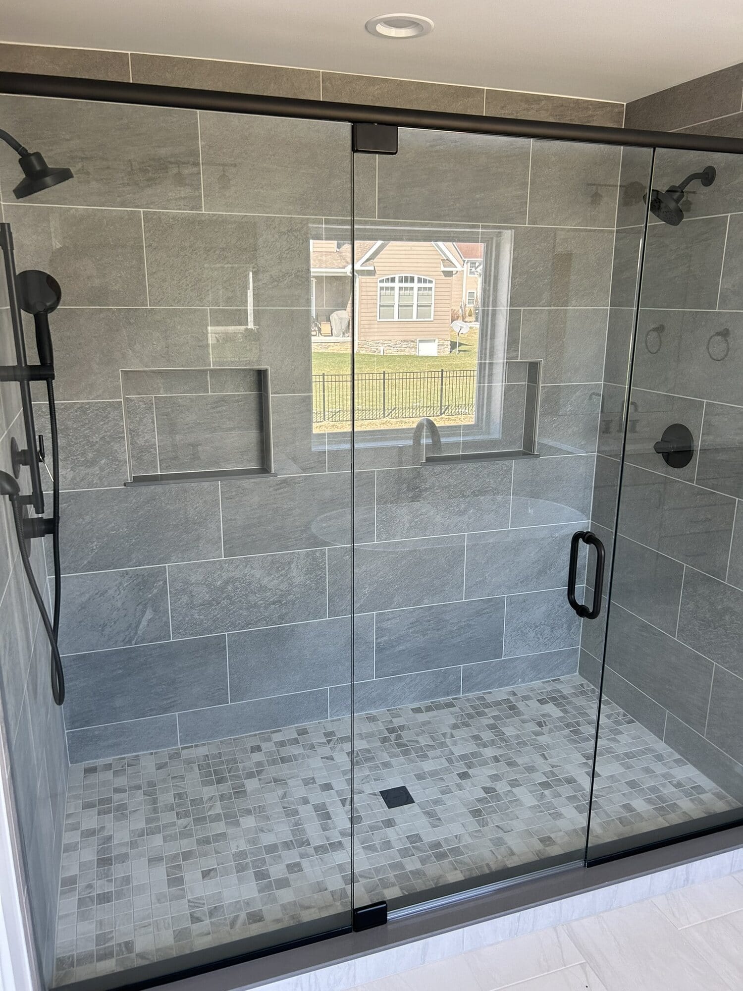 Frameless glass walk-in shower in gray tile with a privacy window, matte black fixtures, and a mosaic tile shower pan.