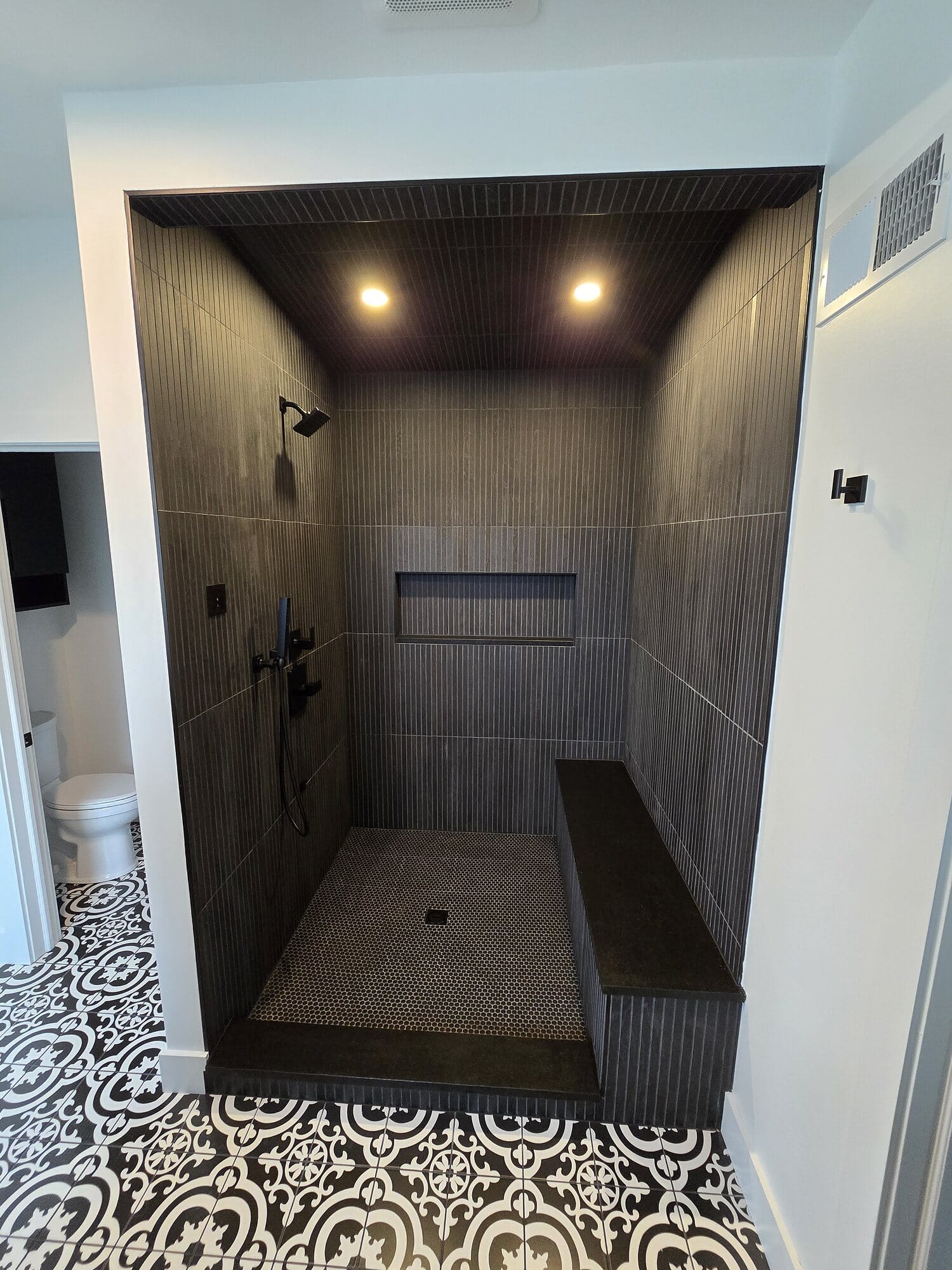 Oversized walk-in shower clad in large-format wood-look tile with a full-width bench, recessed niche, and patterned cement tile floor beyond.