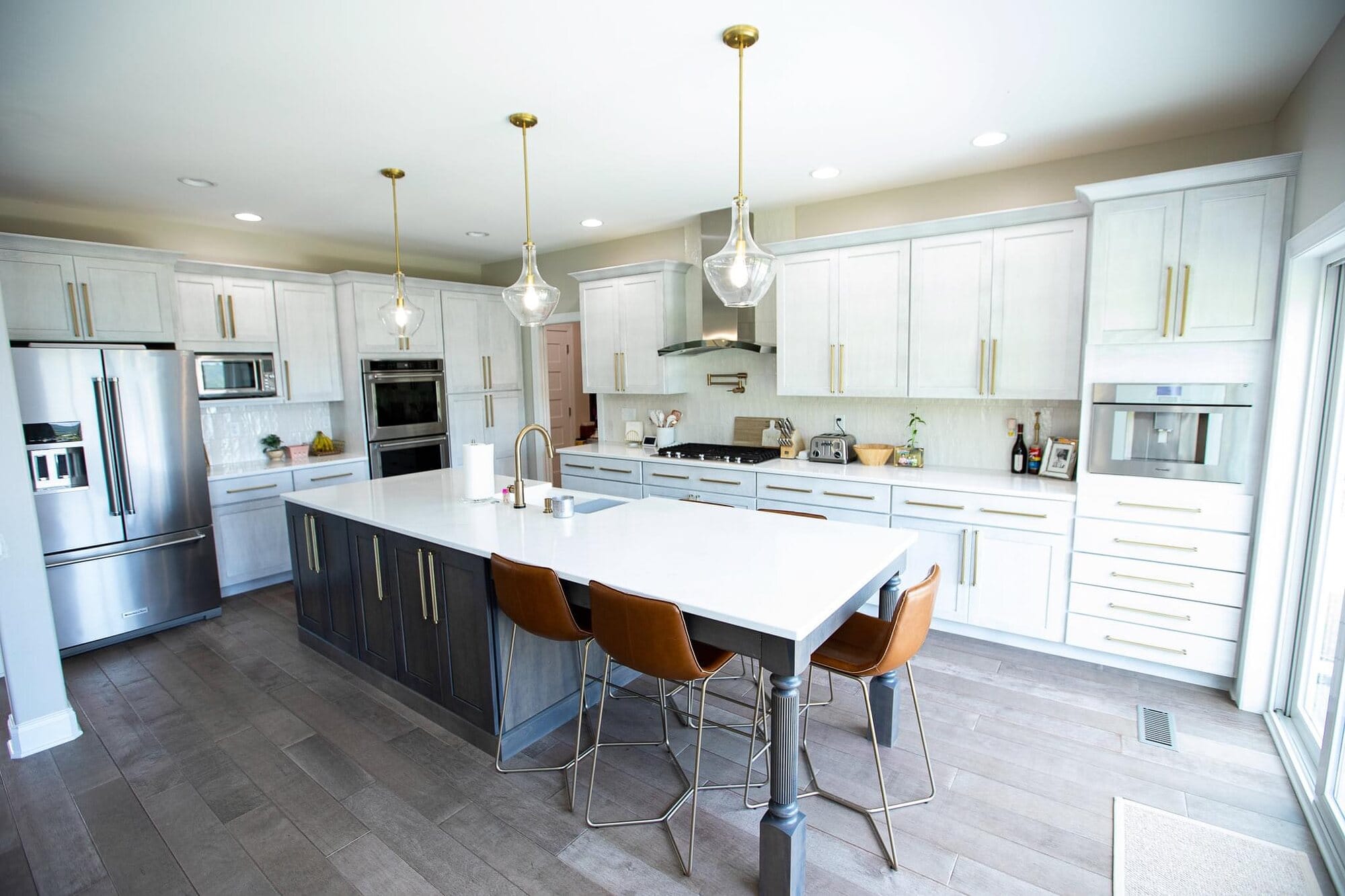 White kitchen with a contrasting dark island, counter seating, glass pendants, wall ovens, and large-format gray tile flooring.