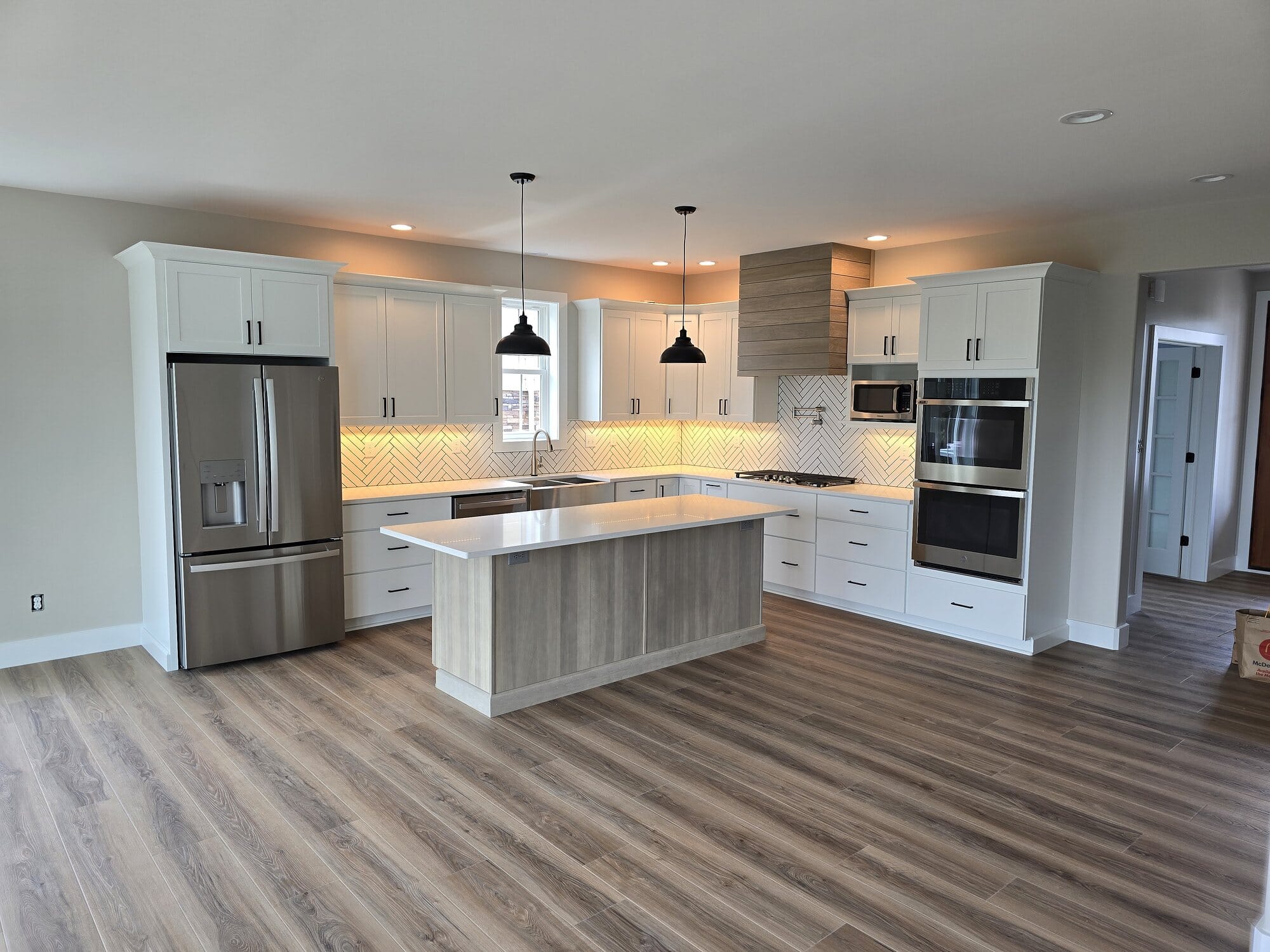 Bright white kitchen with a large center island, stainless steel appliances, pendant lighting, and wood-look plank flooring.