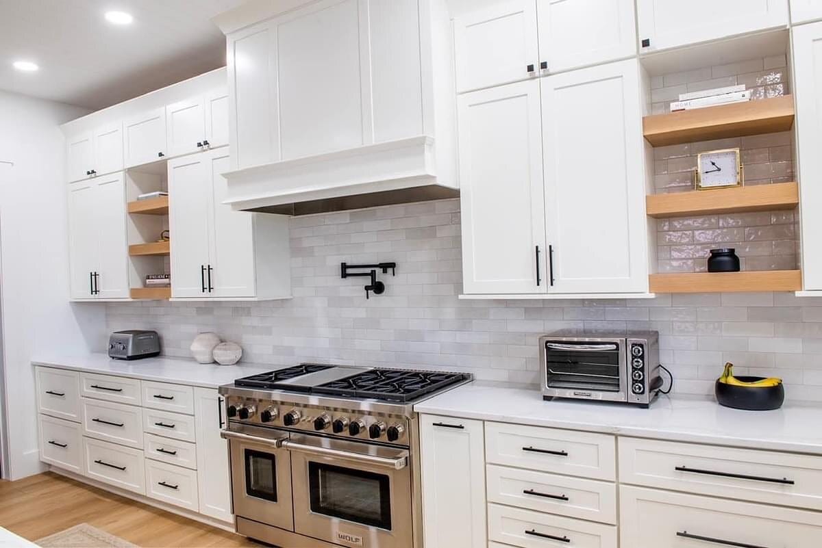 White kitchen range wall with a professional gas range, custom hood surround, pot filler, and floating wood shelves.