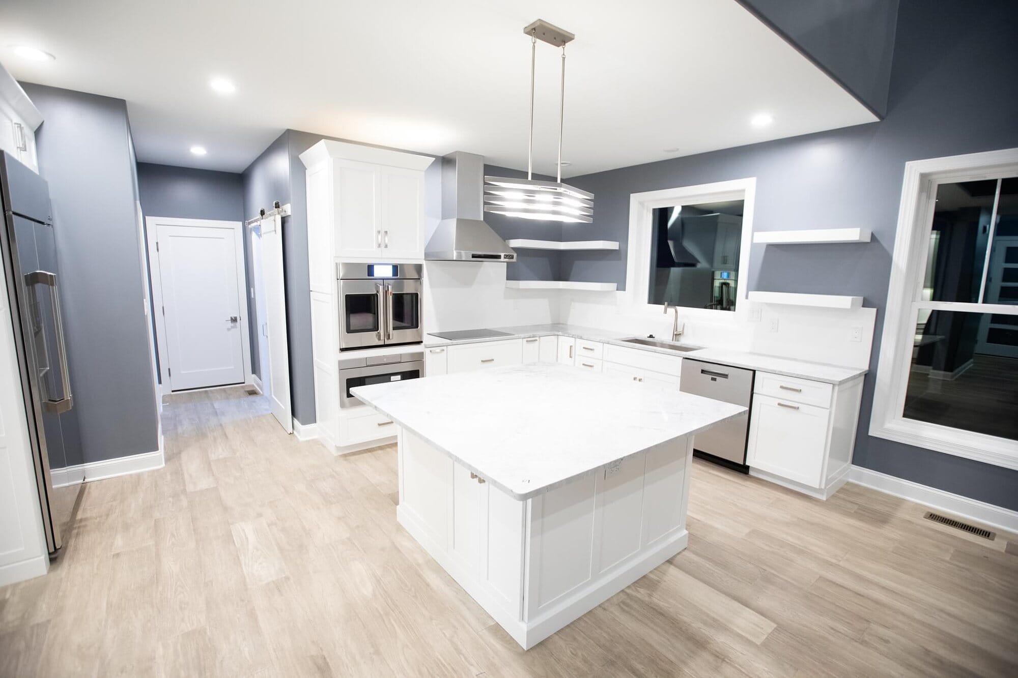 White kitchen with a large square island, quartz countertops, stainless wall ovens, and a stainless vent hood.