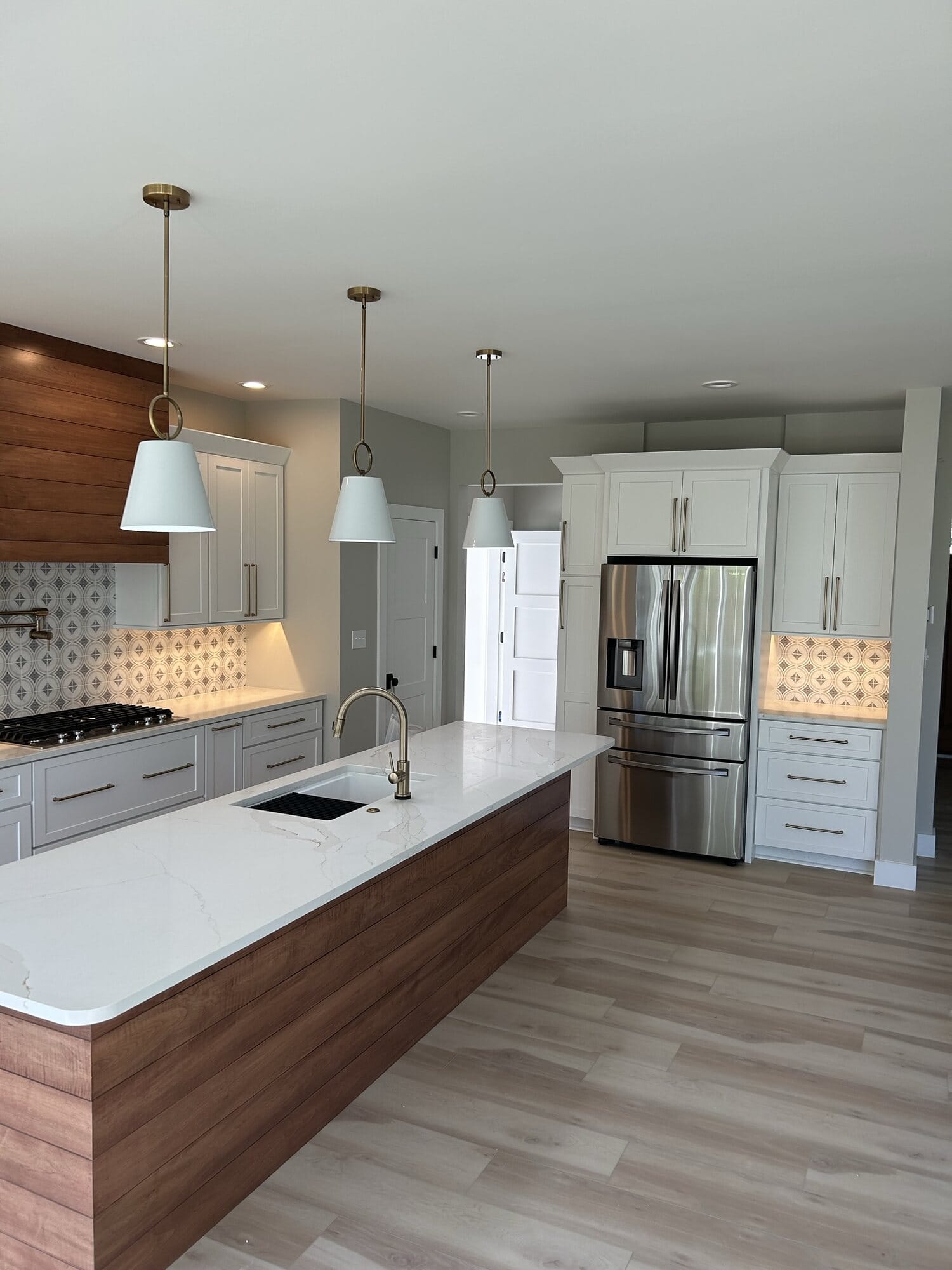 Wide view of a white kitchen with a long walnut island, quartz countertops, stainless refrigerator, and pendant lighting.